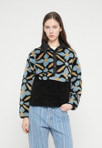 Φλις πουλόβερ με μαύρη βάση και πολύχρωμο ανθικό μοτίβο. Διαθέτει τεταρτημόριο με κουμπιά και υφασμάτινη υφή, συνδυασμένο με ριγωτό τζιν.