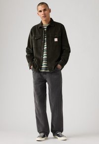 Hombre con pelo corto que lleva una chaqueta de camisa de pana negra sobre una camiseta de rayas verde, pantalones anchos grises y zapatillas negras de corte bajo.
