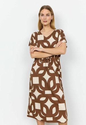 Woman with folded arms wearing a brown and cream geometric patterned knee-length dress with short sleeves and a V-neck.