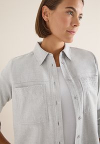 Jasnoszara koszula z guzikami i kołnierzykiem. Posiada dwie kieszenie na piersi ozdobione dekoracyjnymi ćwiekami oraz fakturą materiału.