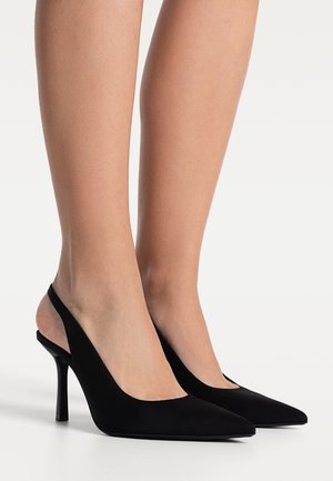 Jambes portant des escarpins slingback noirs à bout pointu avec des talons aiguilles fins sur fond blanc.