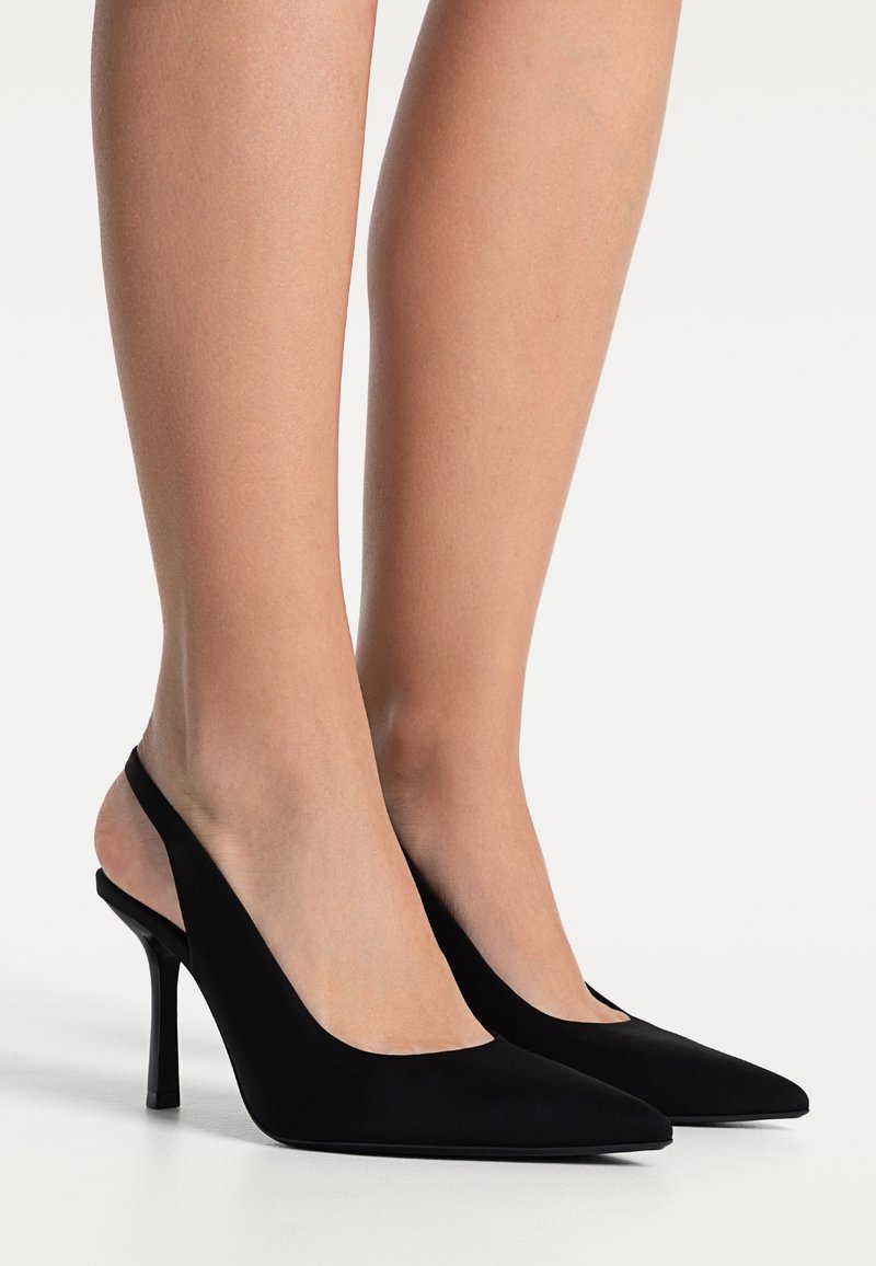 Jambes portant des escarpins slingback noirs à bout pointu avec des talons aiguilles fins sur fond blanc.