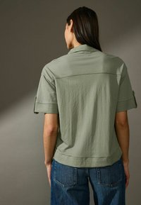 Camicetta verde oliva a maniche corte con vestibilità rilassata, caratterizzata da una cucitura a yoke sulla schiena e polsini arrotolati. Abbinate a jeans in denim blu, di vestibilità ampia.