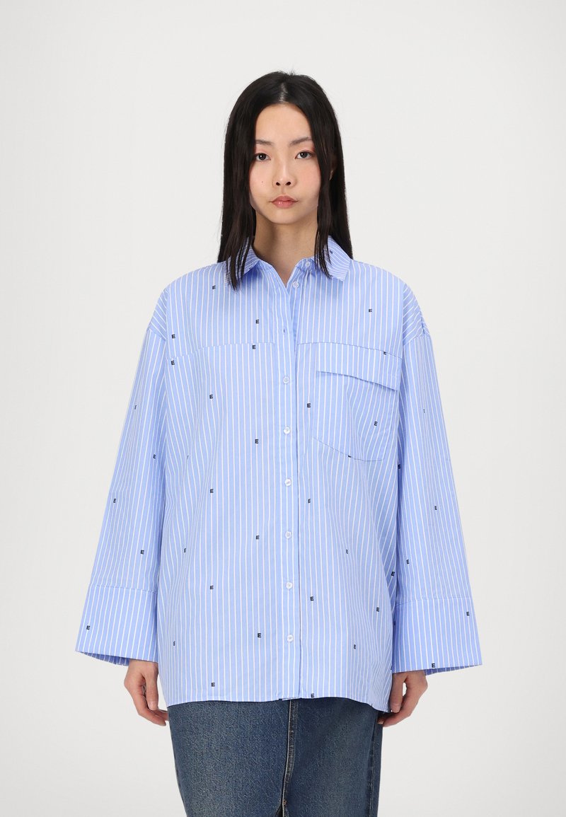 Chemise ample bleu clair à rayures verticales blanches, ornée de petits motifs carrés noirs et d'une poche poitrine ; design à boutons et manches larges.