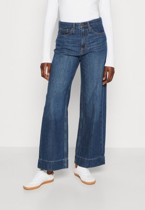 Model trägt weit geschnittene, hoch taillierte blaue Denim-Jeans, weißes Langarmshirt und weiße Sneakers mit Gummisohle vor einfarbigem Hintergrund.