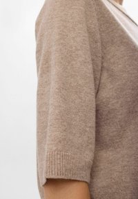 Mjuk beigefärgad stickad kortärmad tröja på en person som visar textur och ribbad mudddetalj mot en vit bakgrund.