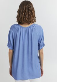 Blusa azzurro chiaro realizzata in un tessuto morbido, con scollatura arricciata e maniche corte elasticizzate, dalla vestibilità ampia e con spacchi laterali.
