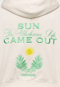 Sweat à capuche beige avec texte brodé vert « SUN The Wholesome Life Came Out », un soleil jaune, deux feuilles de palmier vertes et des lignes ondulées bleues en dessous.
