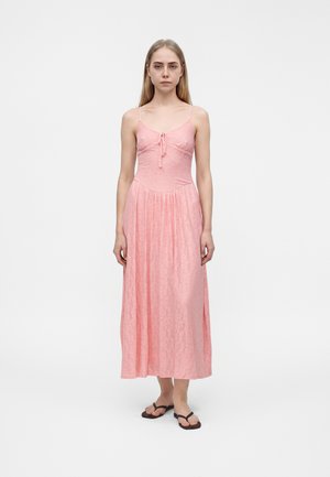 Junge Frau mit langen blonden Haaren, die ein ärmelloses, pinkfarbenes Spitzen-Maxikleid und schwarze Flip-Flops trägt, steht vor einem einfarbigen Hintergrund.
