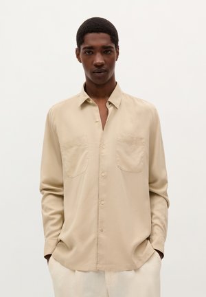 Camisa de manga larga beige hecha de tela suave. Presenta un cierre frontal con botones, dos bolsillos en el pecho y un corte holgado.
