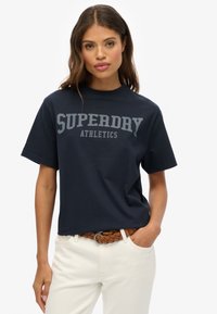 T-shirt en coton bleu marine à manches courtes, arborant un grand logo gris "SUPERDRY ATHLETICS" sur la poitrine.