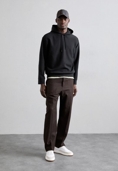 Svart huvtröja med strukturerad yta, bruna randiga byxor och vita sneakers. Kappen har flerfärgade vertikala ränder.
