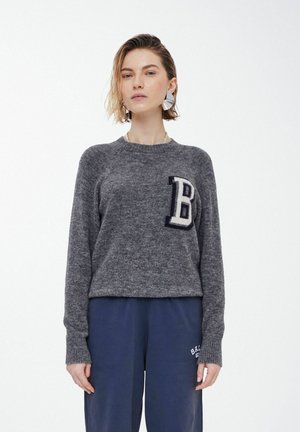 Grå strikket sweater med rund halsudskæring og rib strikkede manchetter, der har et cremefarvet "B"-patch på venstre bryst. Blød tekstur, afslappet pasform.