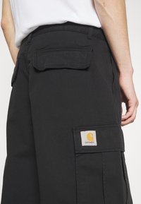 Mustat cargo-housut, jotka on valmistettu kestävästä kankaasta. Niissä on sivutaskut, suorakulmainen takatasku ja Carhartt-logopatchi oranssilla ja valkoisella.