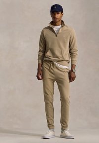 Beiger Pullover mit halbseitigem Reißverschluss und Kängurutasche, kombiniert mit passenden Jogginghosen mit Kordelzug. Dazu ein weißes T-Shirt und Sneakers.