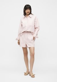 Femme portant une chemise rose clair à manches longues rayée, rentrée dans un short à cordon assorti et des sandales beiges avec des nœuds, debout sur fond blanc.