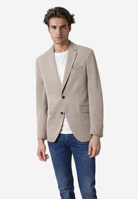 Hellbeiges Blazer aus strukturiertem Stoff, mit zwei Knöpfen und zwei Taschen, kombiniert mit einem weißen T-Shirt und blauen Jeans; lässige Passform.