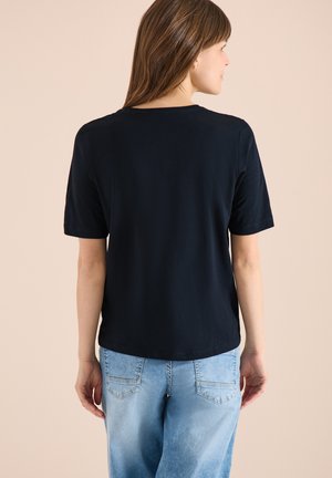 Femme aux cheveux bruns raides portant un t-shirt noir uni à manches courtes et un jean en denim bleu clair, tournée de dos sur un fond beige.