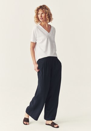 Femme portant un haut blanc à manches courtes avec un col en V, un pantalon noir ample et des sandales foncées, debout les mains dans les poches, regardant de côté.
