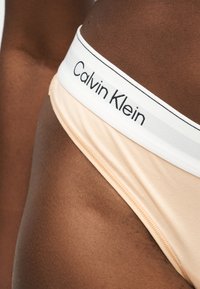 Άτομο που φοράει μπεζ εσώρουχο με λευκή μέση που φέρει την επιγραφή "Calvin Klein" σε μαύρο χρώμα πάνω σε σκούρο δέρμα.