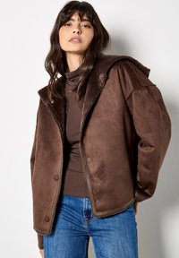 Giacca in finta pelle scamosciata marrone con colletto texturizzato e morbido, chiusura a bottoni e vestibilità oversize, abbinata a una maglia marrone e jeans blu.