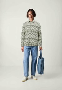 Grün gemustertes Langarmhemd, hellblaue Jeans, weiße Sneakers mit blauen Streifen und eine blaue Tragetasche.
