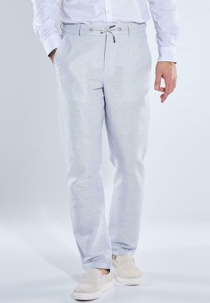 Mann trägt hellgraue Hose mit Kordelzug und weiße Slip-On-Schuhe, steht mit entspannt an den Seiten hängenden Händen vor einfarbigem Hintergrund.