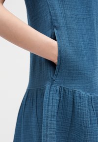 Χέρι που έχει εισαχθεί στην πλαϊνή τσέπη ενός υφασμάτινου μπλε φορέματος με σούρωμα στη μέση, αναδεικνύοντας λεπτομέρειες του υφάσματος και του σχεδιασμού της τσέπης.