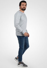 Grauer Sweatshirt mit langen Ärmeln, rundem Ausschnitt und gerippten Bündchen, kombiniert mit blauen Jeans und grauen Sportschuhen mit weißen Streifen.