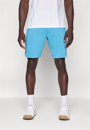 Mann trägt hellblaue Sportshorts, weiße Sportschuhe, weiße Socken und ein weißes Oberteil und steht vor einem einfarbigen Hintergrund.
