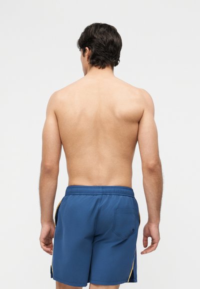 Mężczyzna o ciemnych włosach stoi tyłem, bez koszulki, w niebieskich kąpielówkach z żółtymi paskami po bokach, na jednolitym jasnym tle.