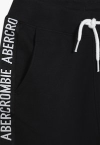 Zwarte joggingbroek met een witte trekkoord en zijpanelen met herhaalde tekst. De stof is glad met een lichte glans.