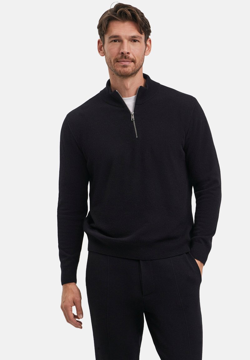 Schwarzer Strickpullover mit hohem Kragen und halber Reißverschlussöffnung. Glatte Textur, lange Ärmel und taillierte Passform, kombiniert mit dunklen Hosen.