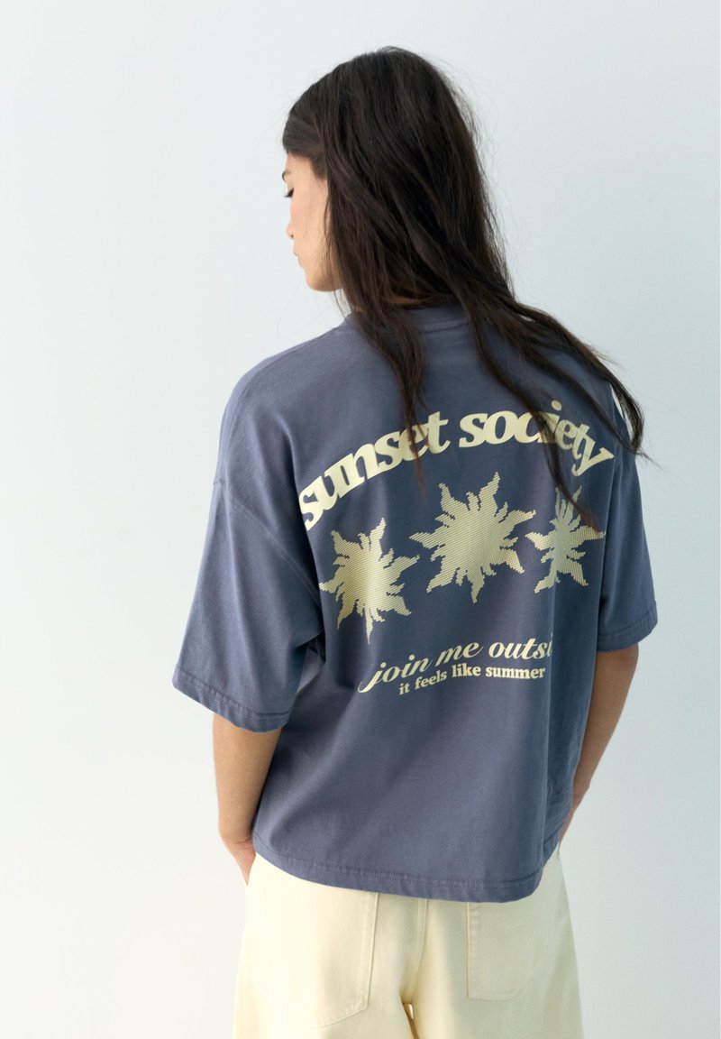 Žena s dlhými tmavými vlasmi, ktorá má na sebe modré nadrozmerné tričko s žltými slnečnými grafikami a textom „sunset society“ a „join me outside“.