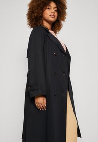 Navy dobbeltspent trench coat med store knapper, brede revers og lange ermer; har sidesplitt for økt bevegelsesfrihet.