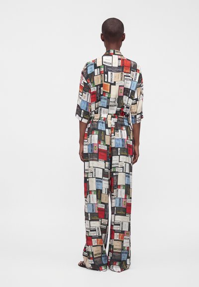 Personne portant une combinaison ample avec des motifs rectangulaires colorés d'étiquettes et des manches courtes, debout de dos sur un fond uni.