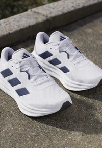 White athletic shoes with navy blue stripes and white laces, placed on a textured concrete surface near a stone curb.