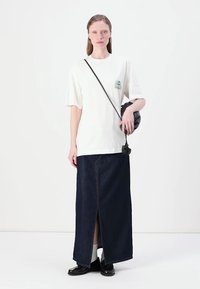 White oversized t-shirt with a green graphic, paired with a dark denim maxi skirt featuring a front slit. Black crossbody bag and shoes.