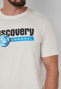Weißes Baumwoll-T-Shirt mit "Discovery CHANNEL" in fetten schwarzen Buchstaben, begleitet von einem blauen Akzentstreifen und einer blauen Planeten-Grafik. Kurze Ärmel.