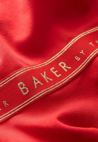 Červená látka s hladkou textúrou má výraznú zlatú nálepku s nápisom "BAKER" na kontrastnej stužke. Jednoduchý a elegantný dizajn.