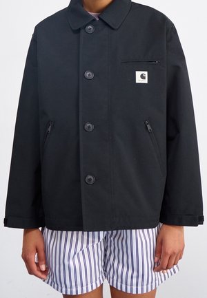 Persoon draagt een zwart jasje met knopen en borst- en zijzakken met rits, gecombineerd met witte shorts met verticale zwarte strepen.