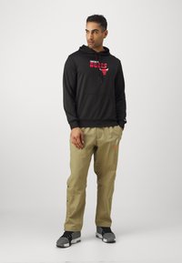 Sweatshirt noir avec le logo "Chicago Bulls" rouge ; pantalon beige avec des boutons sur les côtés et le logo Nike ; chaussures de sport grises ; arrière-plan neutre.