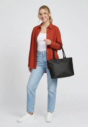 Smilende kvinde iført rustfarvet skjorte med knapper, hvid tanktop, lyseblå jeans, hvide sneakers, der holder en stor tekstureret sort shopper ved håndtagene.