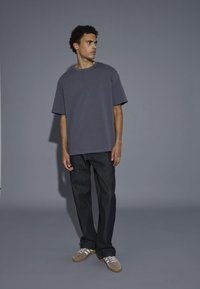 T-shirt oversized grigio ardesia a maniche corte abbinato a jeans denim scuri. Il modello indossa sneakers in suede marroni con strisce bianche.