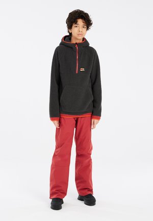 Pull en polaire gris foncé avec des accents rouges, fermeture éclair avant mi-hauteur, capuche et poche avant, associé à un pantalon rouge imperméable et des chaussures noires.