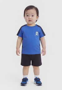 Παιδάκι που στέκεται, φοράει μπλε και μαύρο κοντομάνικο μπλουζάκι με λογότυπο, μαύρο σορτς, λευκές κάλτσες και μπλε αθλητικά παπούτσια σε λευκό φόντο.