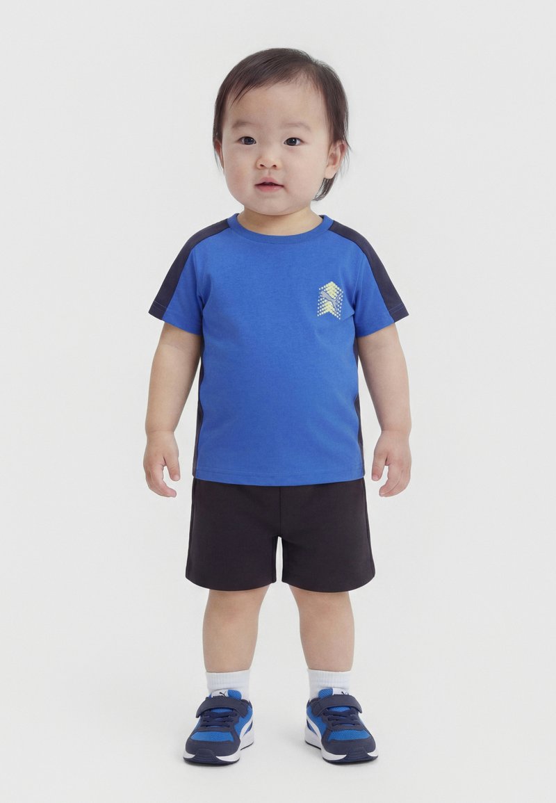 Παιδάκι που στέκεται, φοράει μπλε και μαύρο κοντομάνικο μπλουζάκι με λογότυπο, μαύρο σορτς, λευκές κάλτσες και μπλε αθλητικά παπούτσια σε λευκό φόντο.