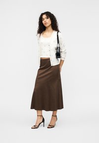 Ελαφρύ κρεμ ζακέτα με διακριτικό σχέδιο, φορεμένο πάνω από λευκό τοπ, συνδυασμένο με καφέ μίντι φούστα και μαύρα πέδιλα με λουράκια και τακούνι.