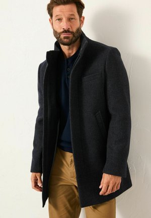 Dunkelgrauer Wollmischmantel mit hohem Kragen, seitlichen Taschen und geradem Schnitt, getragen über einem marineblauen Hemd und khakifarbenen Hosen.