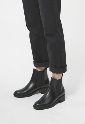 Stivali Chelsea in pelle nera con pannelli elasticizzati ai lati, tacco basso a blocco, punta affilata e texture liscia. Indossati con pantaloni neri arrotolati.